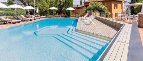Piscina con sillas de descanso y sombrillas, rodeada de árboles y un edificio.