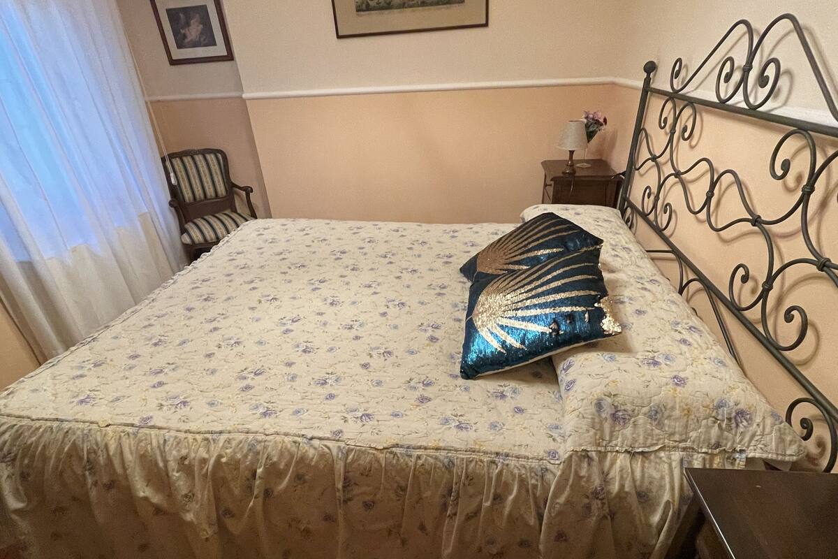 A bedroom with a bed, a chair, a nightstand, a framed picture, and a curtain.