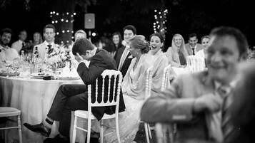 A wedding reception with guests seated at round tables, decorated with floral centerpieces and glassware.