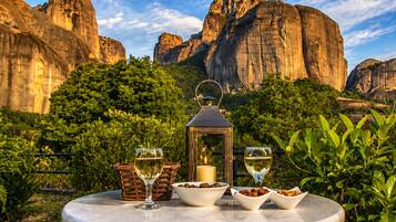 A round table with a candle, wine glasses, and bowls of nuts, set against a backdrop of rocky cliffs and greenery.