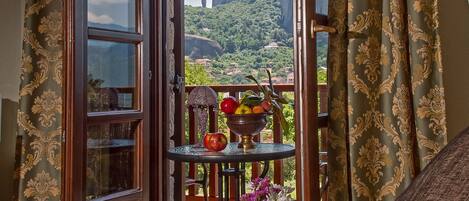 A balcony with a table holding fruit and flowers, a view of a mountainous landscape, and a room with patterned curtains and a bed.