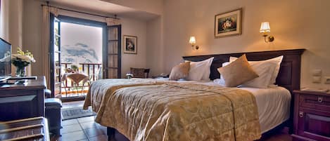 A hotel room with a large bed, a desk with a chair, a view of the mountains through an open door, and two wall-mounted lamps.