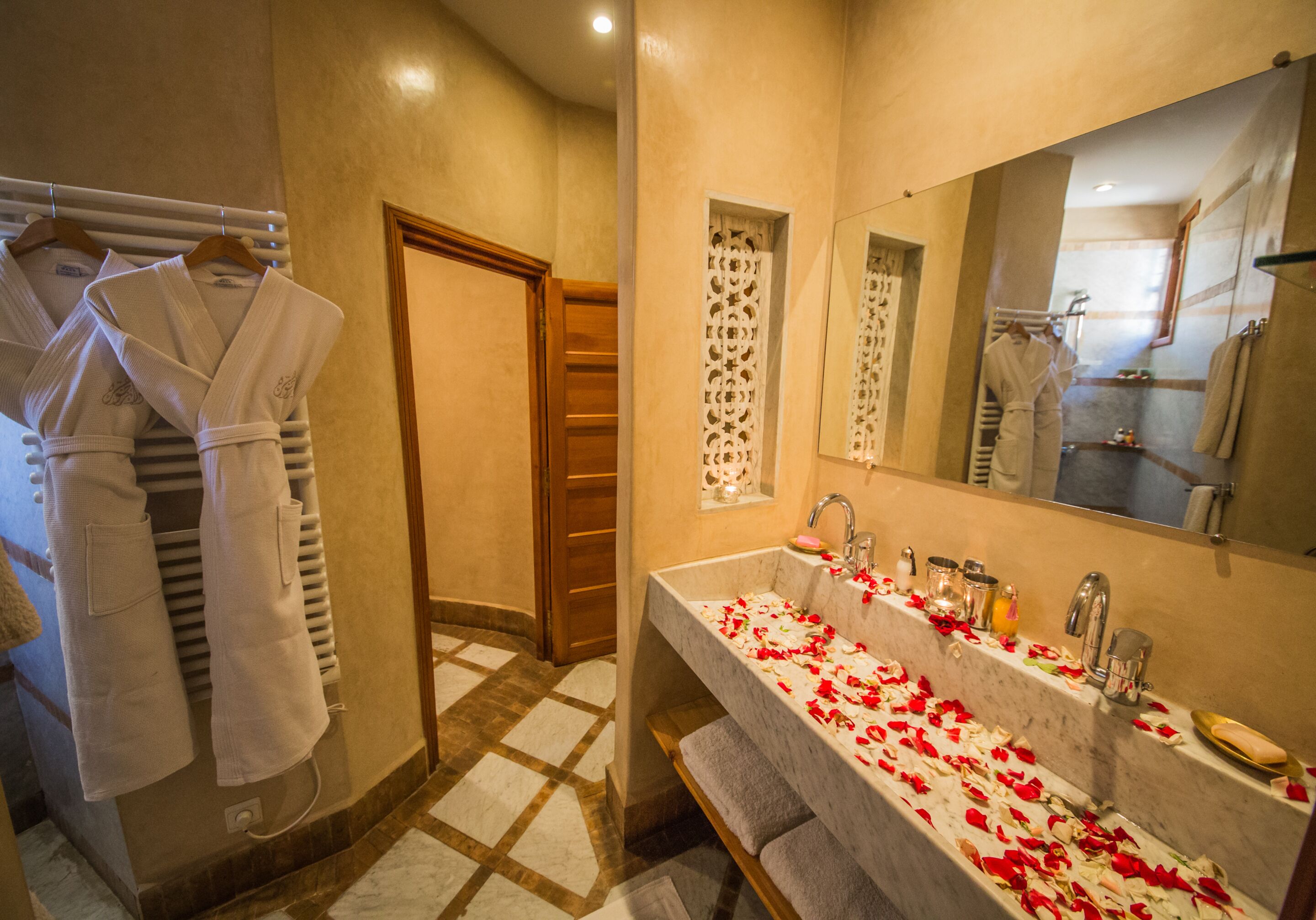 Un baño con un espejo grande, un lavamanos decorado con pétalos de rosa y toallas colgadas en un perchero.