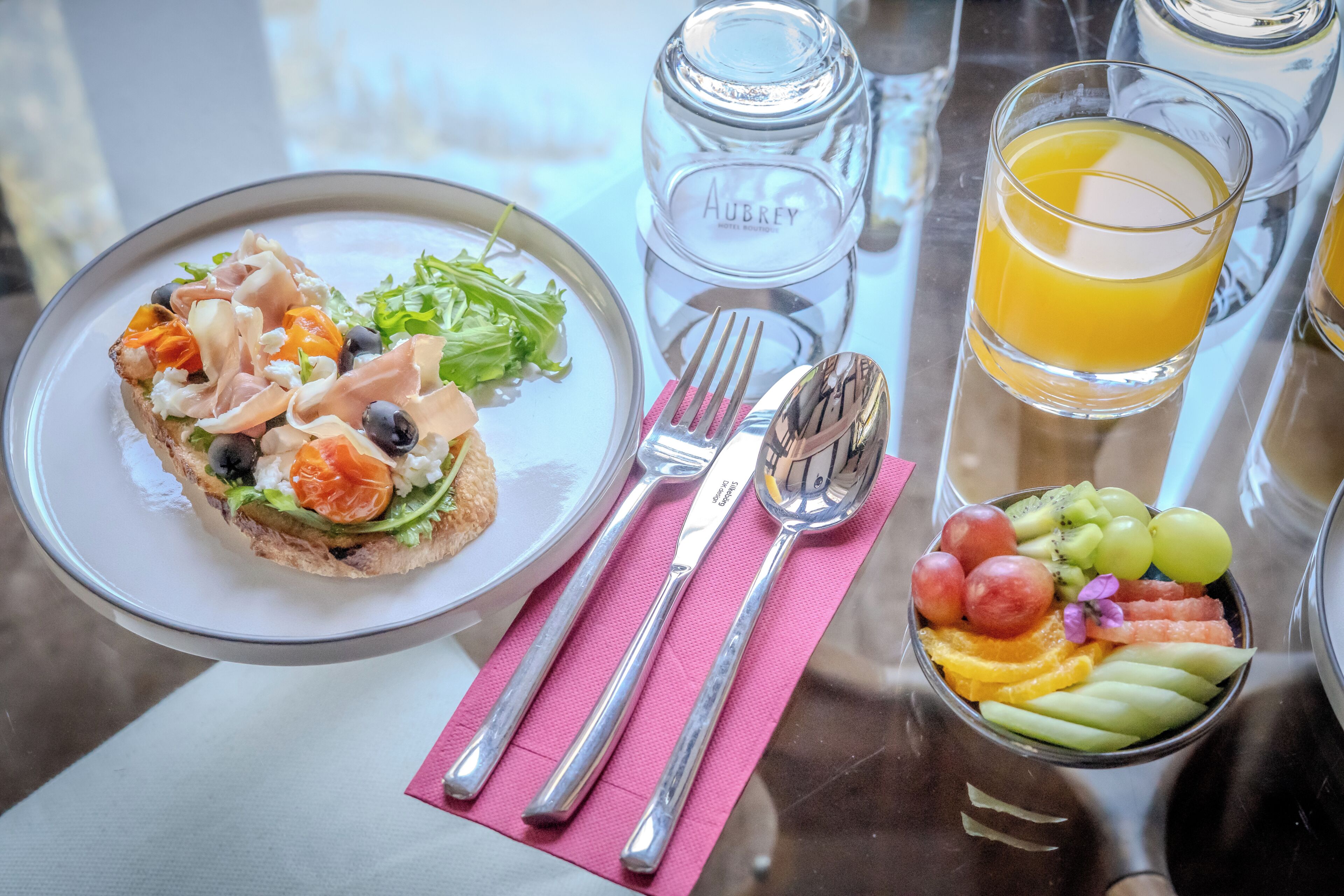 Una mesa con un plato de comida, un vaso de jugo de naranja, un tazón de fruta y una botella etiquetada 'Aubrey'.