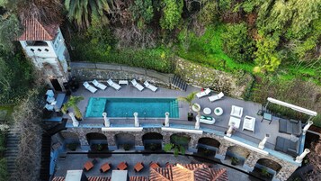 Vista aérea de un complejo turístico con piscina, sillas de descanso y un puente de piedra.