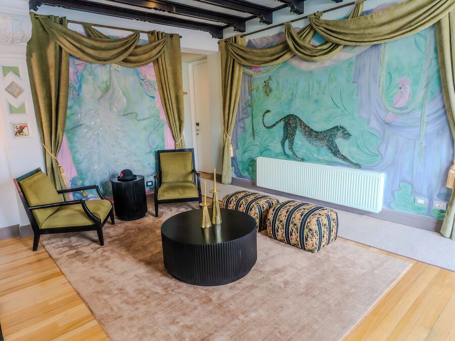 Ein Zimmer mit einem runden schwarzen Tisch, zwei grünen Sesseln, gemusterten Kissen und einem Wandbild mit einem Leoparden, sowie einer Heizung.