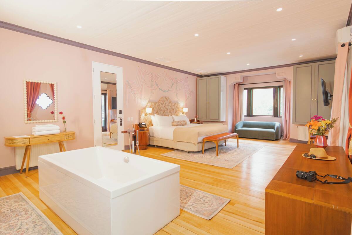 A bedroom with a large white bathtub, a bed with a headboard, a bench, a vanity with a mirror, and a window with curtains.