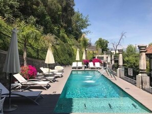 Un área de piscina con sillas de descanso y sombrillas, rodeada de follaje y pilares de piedra.