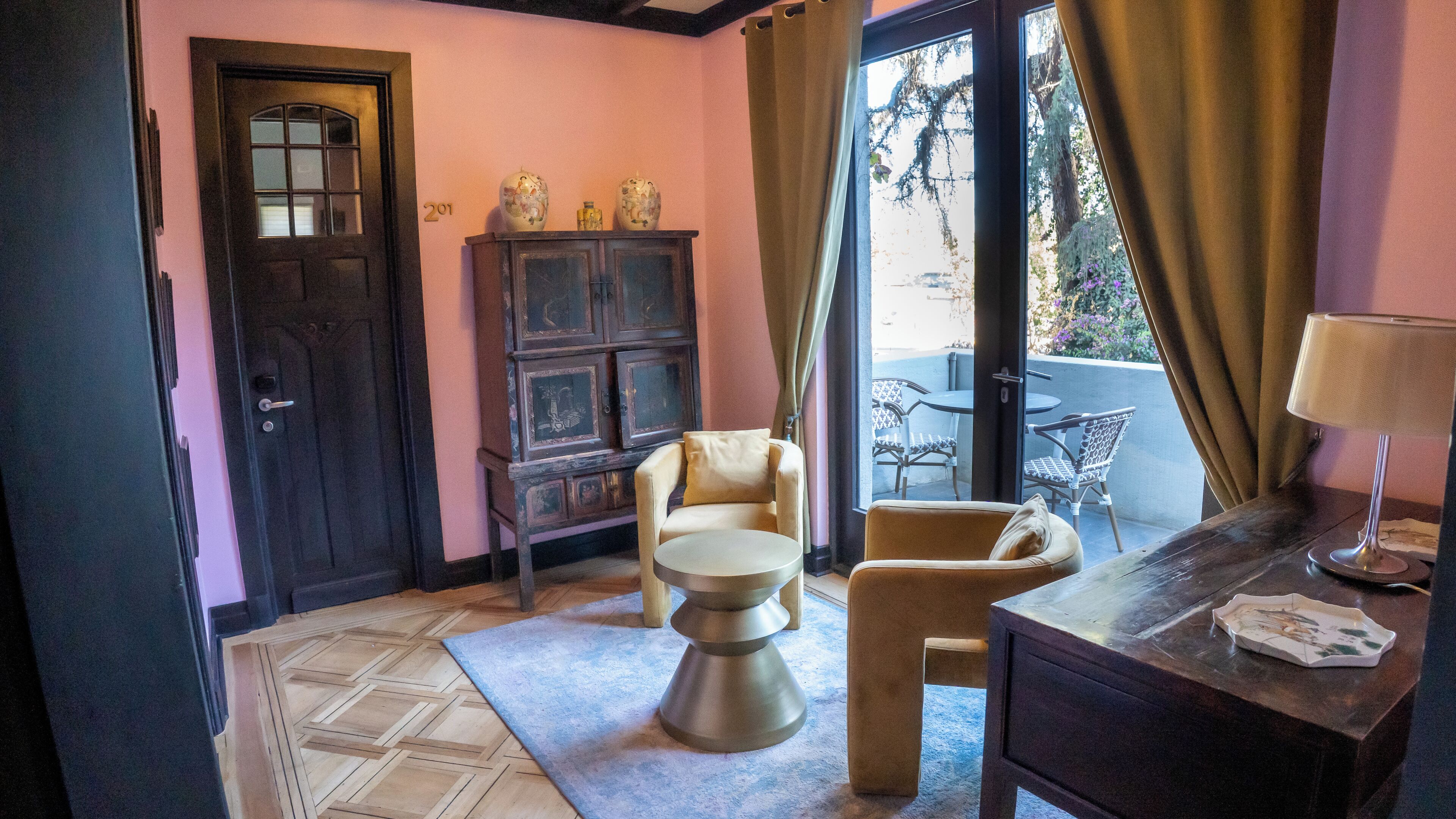 A room with a wooden door, a cabinet with decorative items, a chair, a small table, and a lamp on a wooden dresser.
