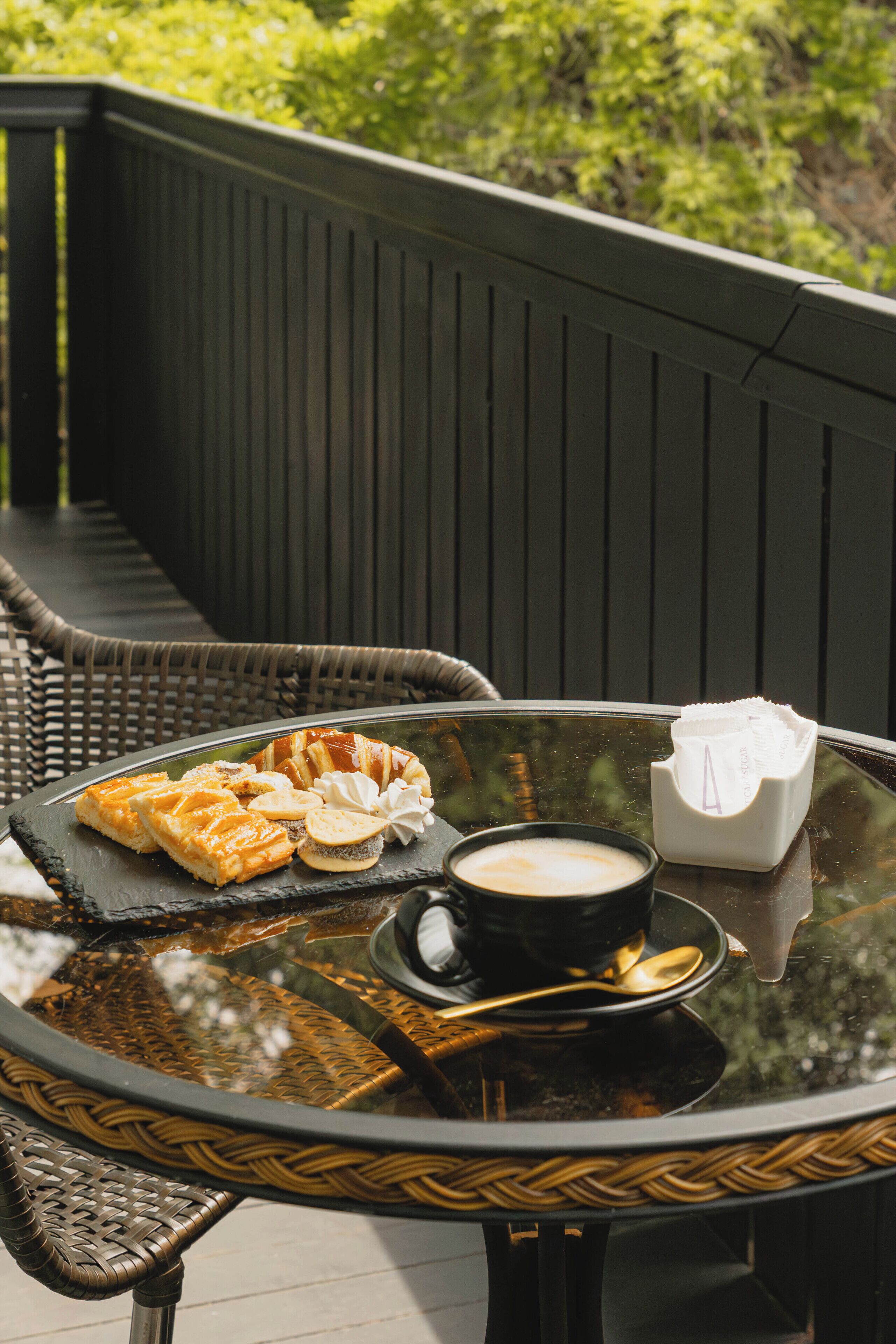 Una mesa de vidrio con pasteles, una taza de café y un sobre de azúcar en un balcón.