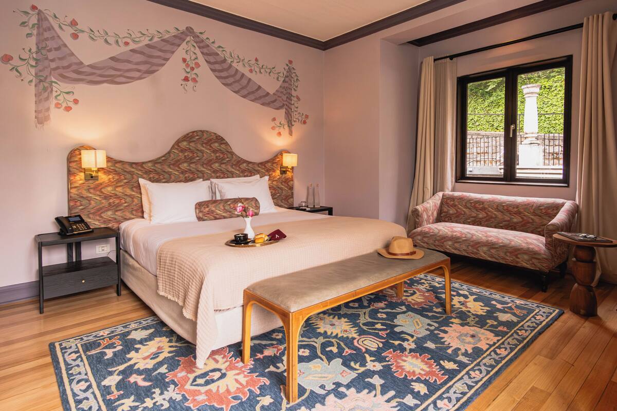 A hotel room with a bed, a bench, a chair, a nightstand, and a window with curtains.