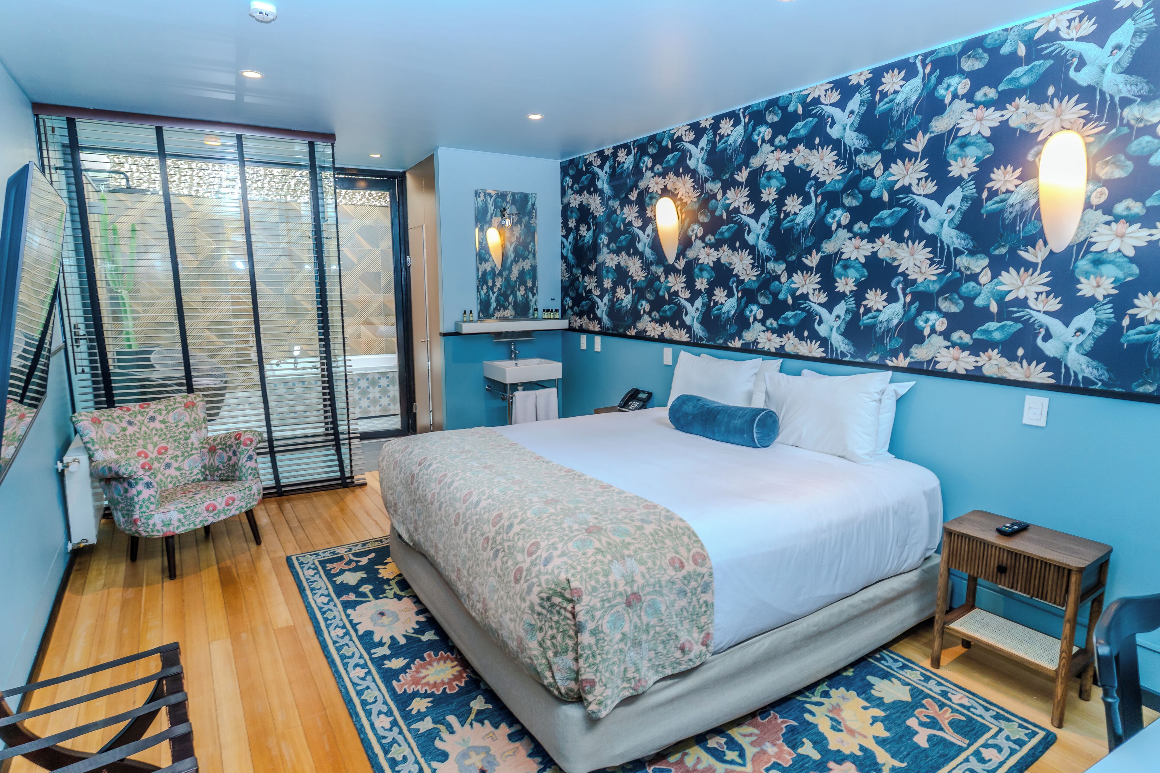 A bedroom with a large bed, a chair, a small table, a mirror, and a wall with a floral pattern.