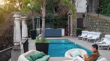 Un área junto a la piscina con sillas de descanso, un edificio blanco con tejado de baldosas y una pared de piedra.