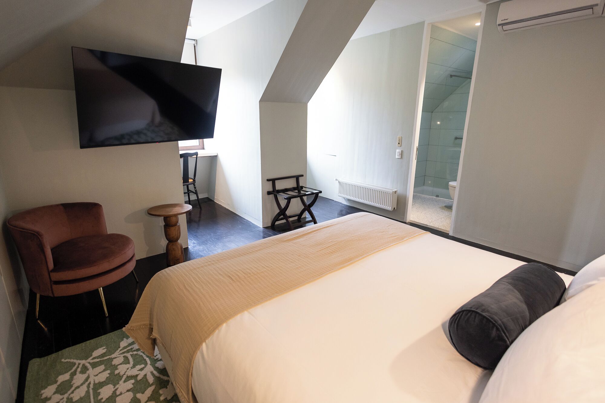 Un dormitorio con una cama grande, un televisor montado en la pared, una silla, una mesita y un cerramiento de vidrio corredera que da al baño.