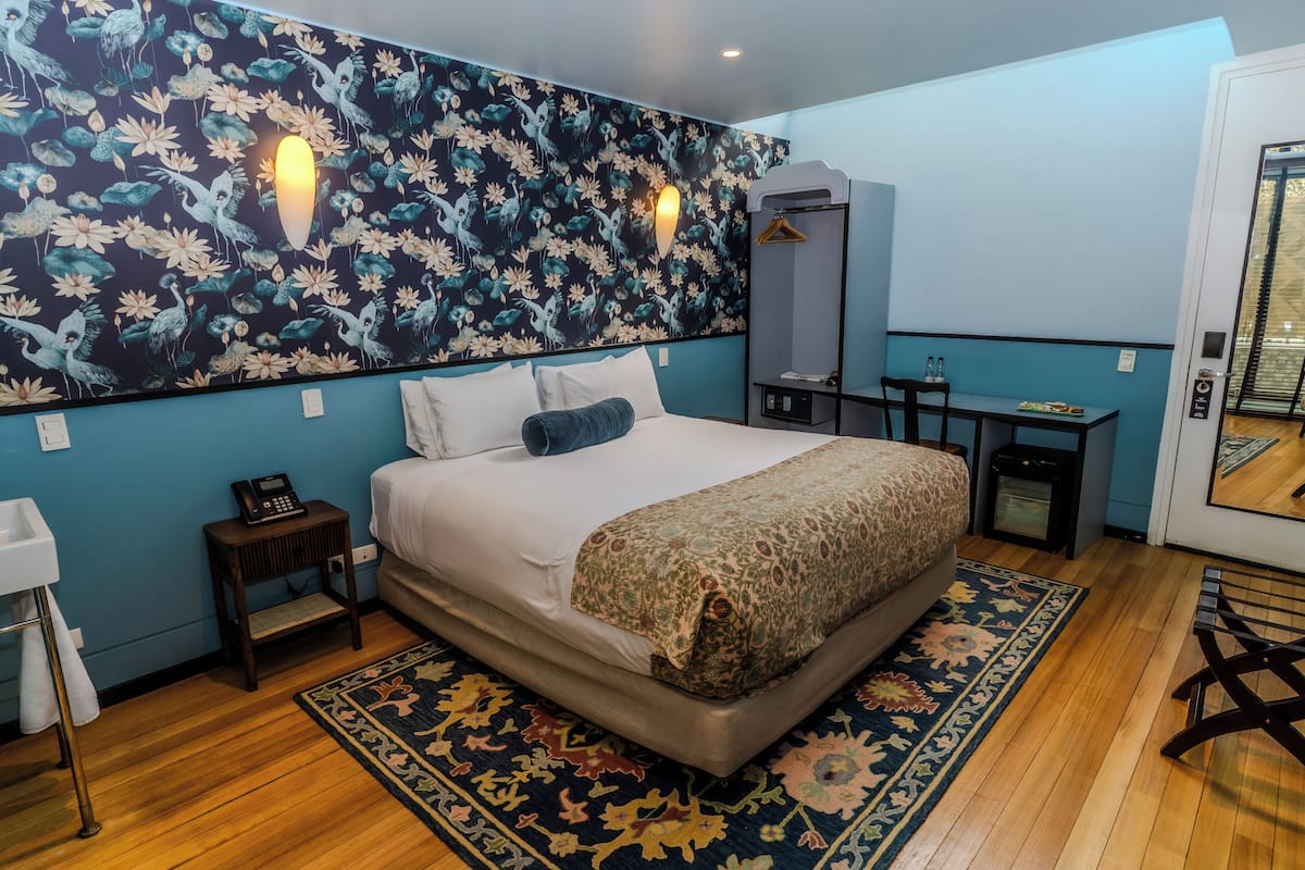 A hotel room with a large bed, a bedside table with a phone, a small table with a chair, a mirror, and a door with a view.
