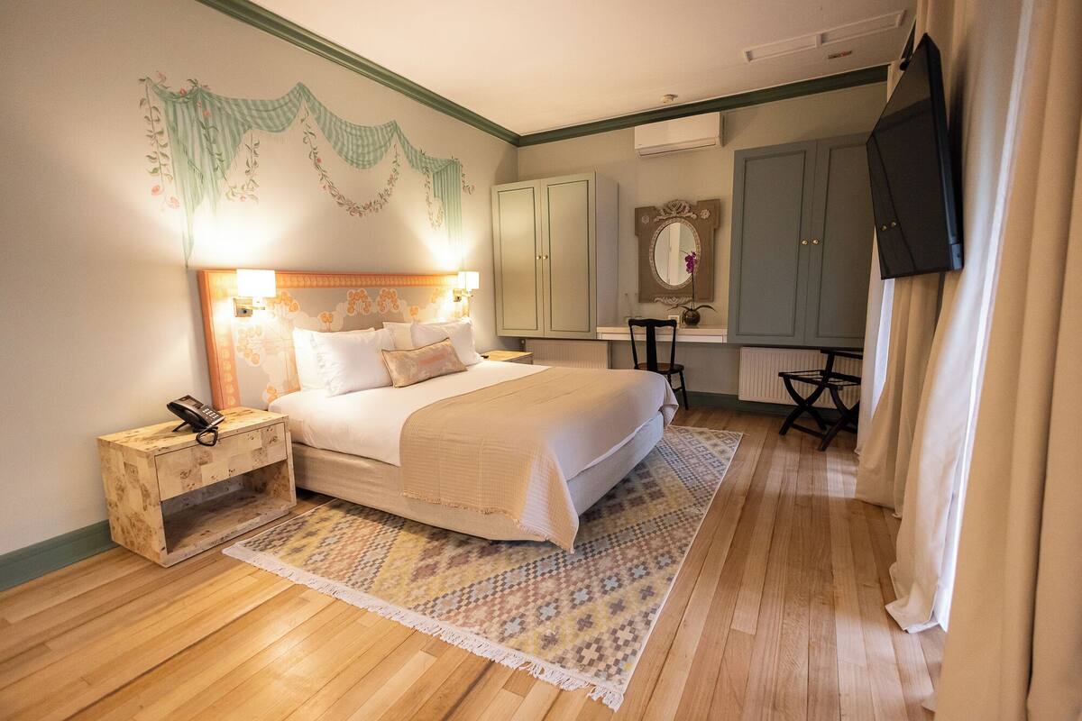 A hotel room with a bed, bedside table with a phone, a desk with a chair, a mirror, and a television.