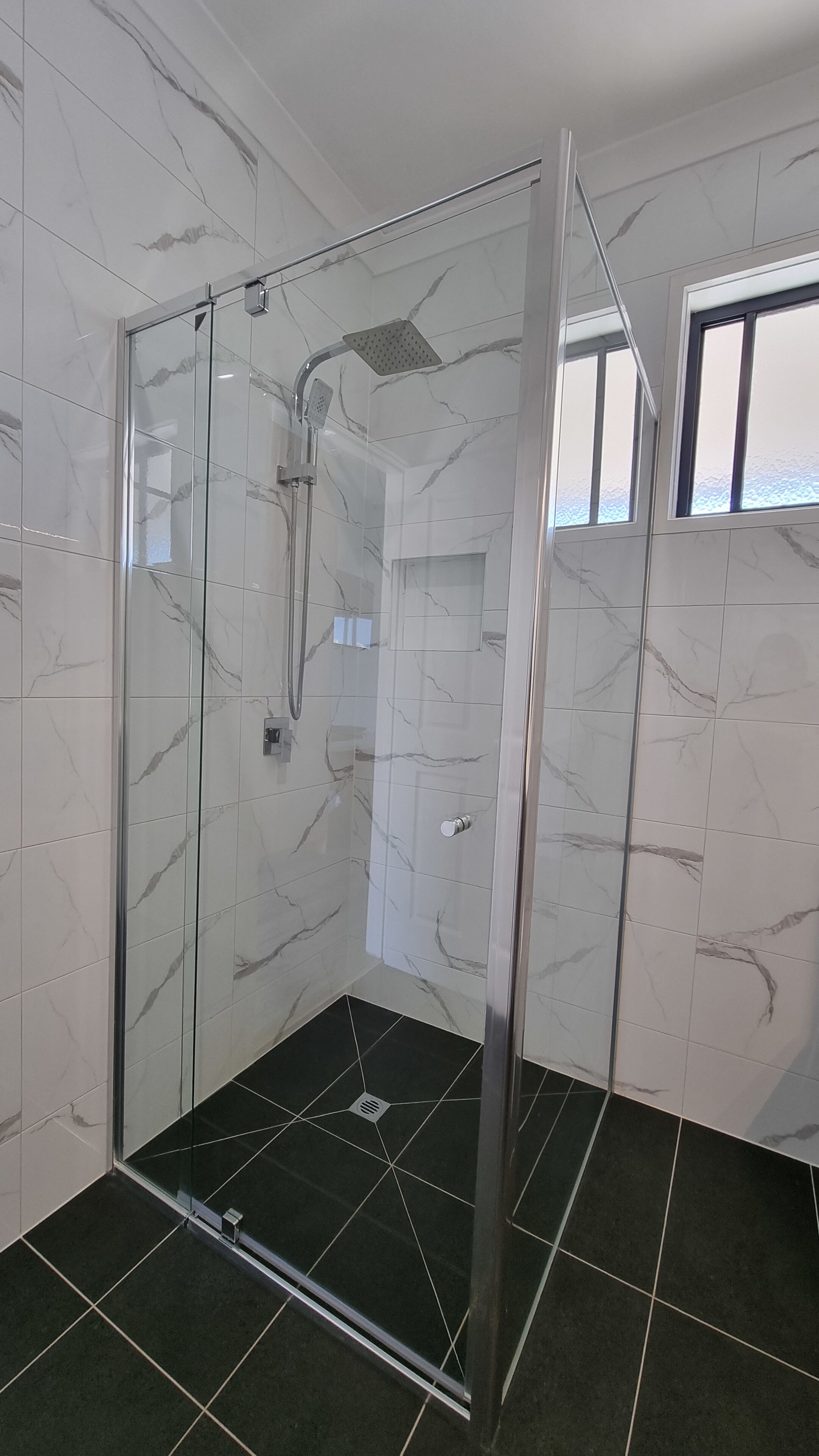 A glass-enclosed shower with a handheld showerhead, a control panel, and a black tiled floor.