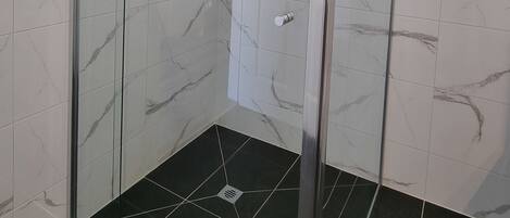 A glass-enclosed shower with a handheld showerhead, a control panel, and a black tiled floor.