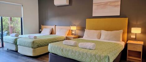 Habitación de hotel con dos camas, cada una con una colcha verde estampada y almohadas blancas, un ventanal grande y un aire acondicionado de pared.