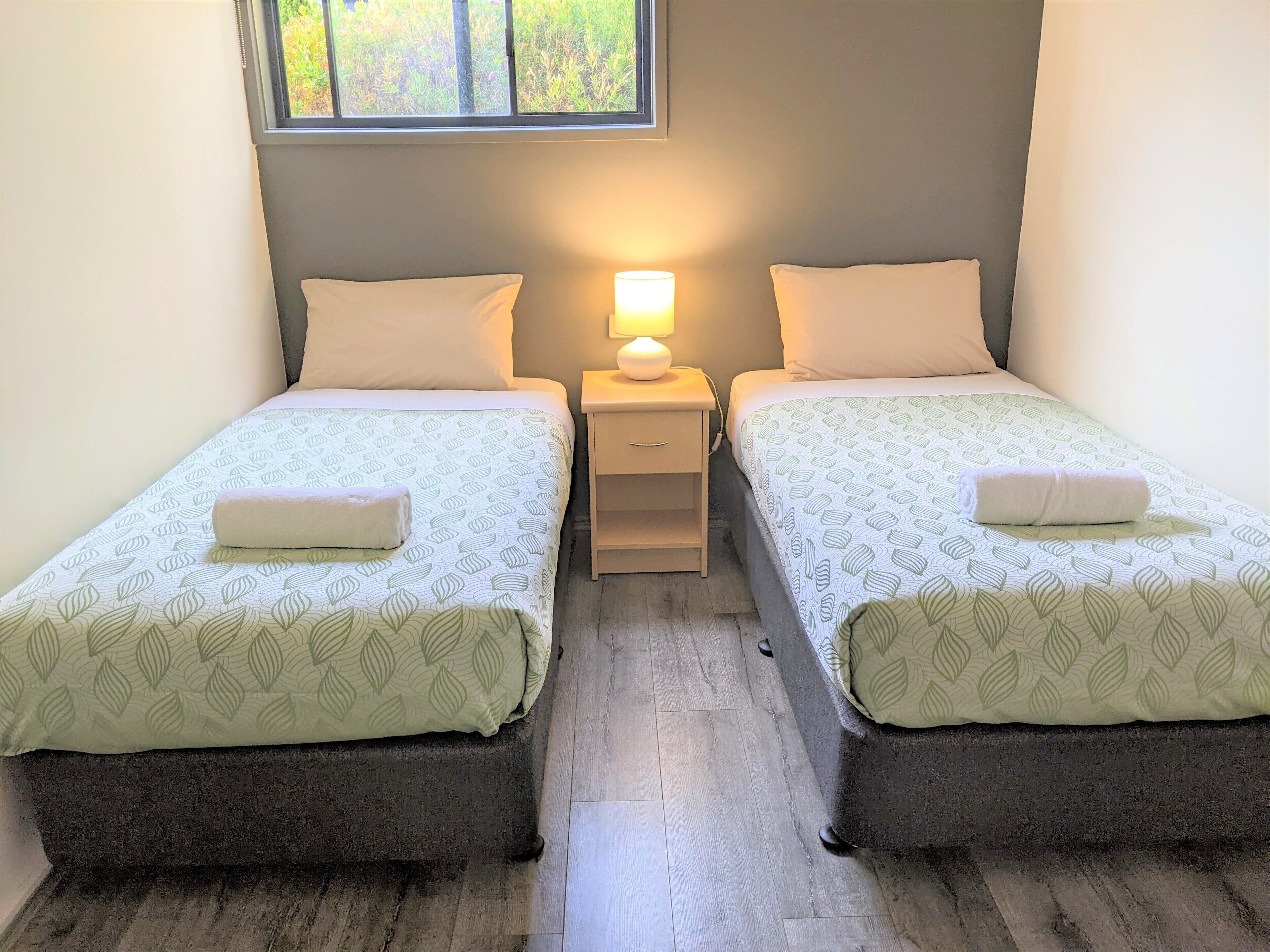 Una habitación con dos camas individuales, cada una con una almohada y una toalla enrollada. Hay una mesita de noche con una lámpara entre las camas.