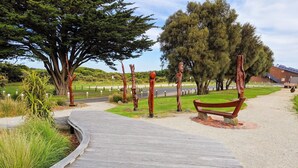 A wooden boardwalk leading to a bench in a park with trees and a building in the background. - Seaview Motel and Apartments (Apollo Bay)