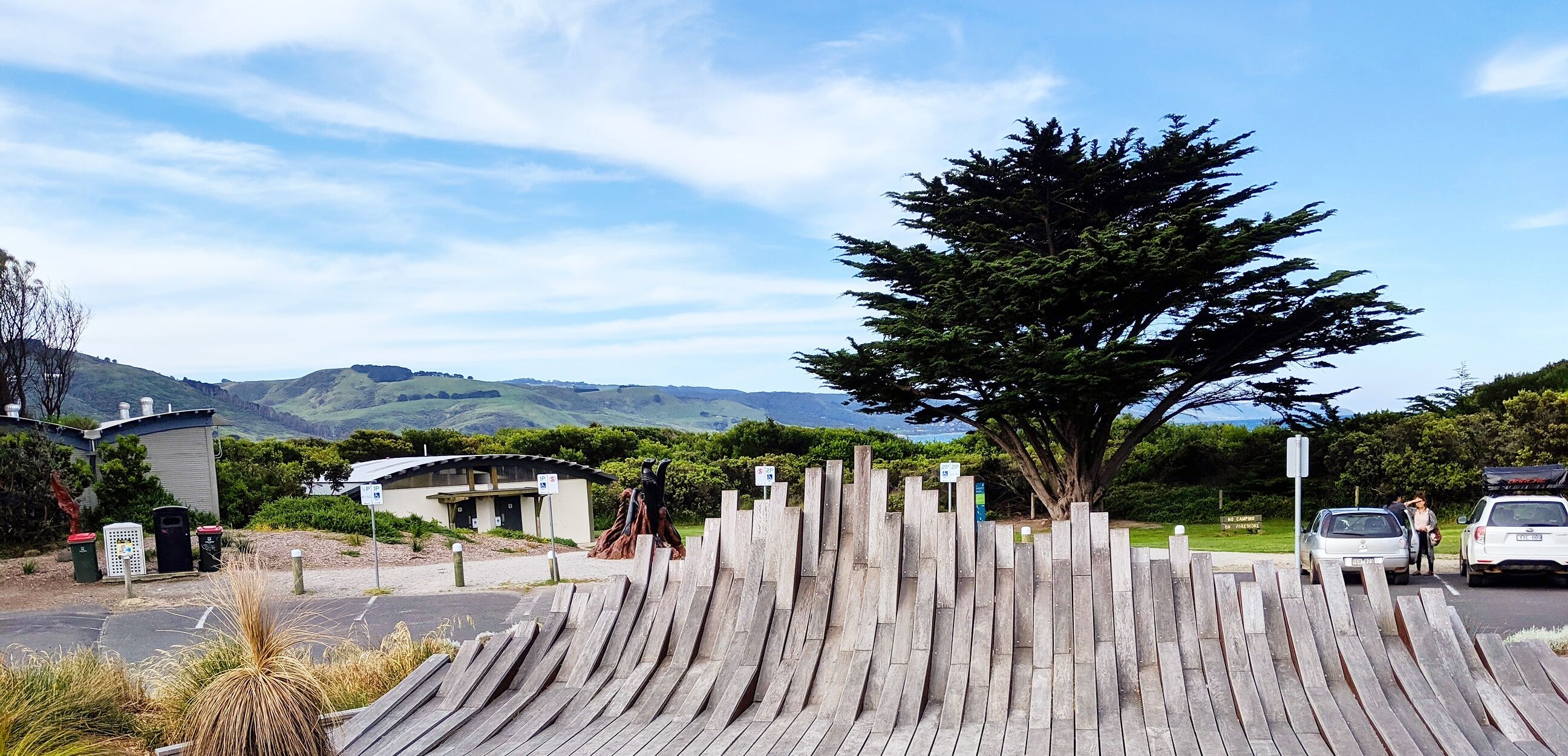 A wooden fence with a tree in the center, a building with a curved roof, and a parking area with cars.