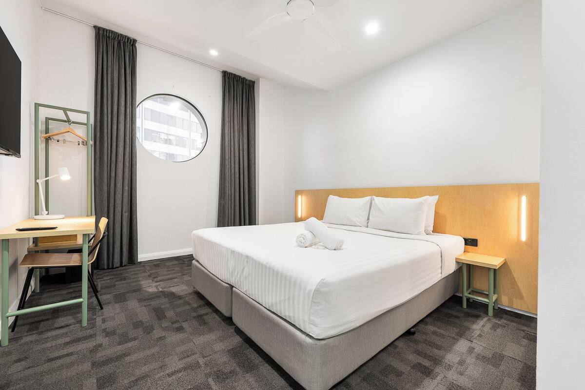 Habitación de hotel con cama, escritorio, silla, espejo redondo y mesita de noche.