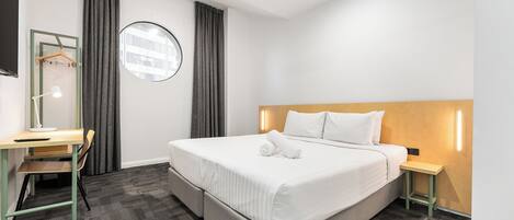 A hotel room with a bed, a desk, a chair, a round mirror, and a nightstand.
