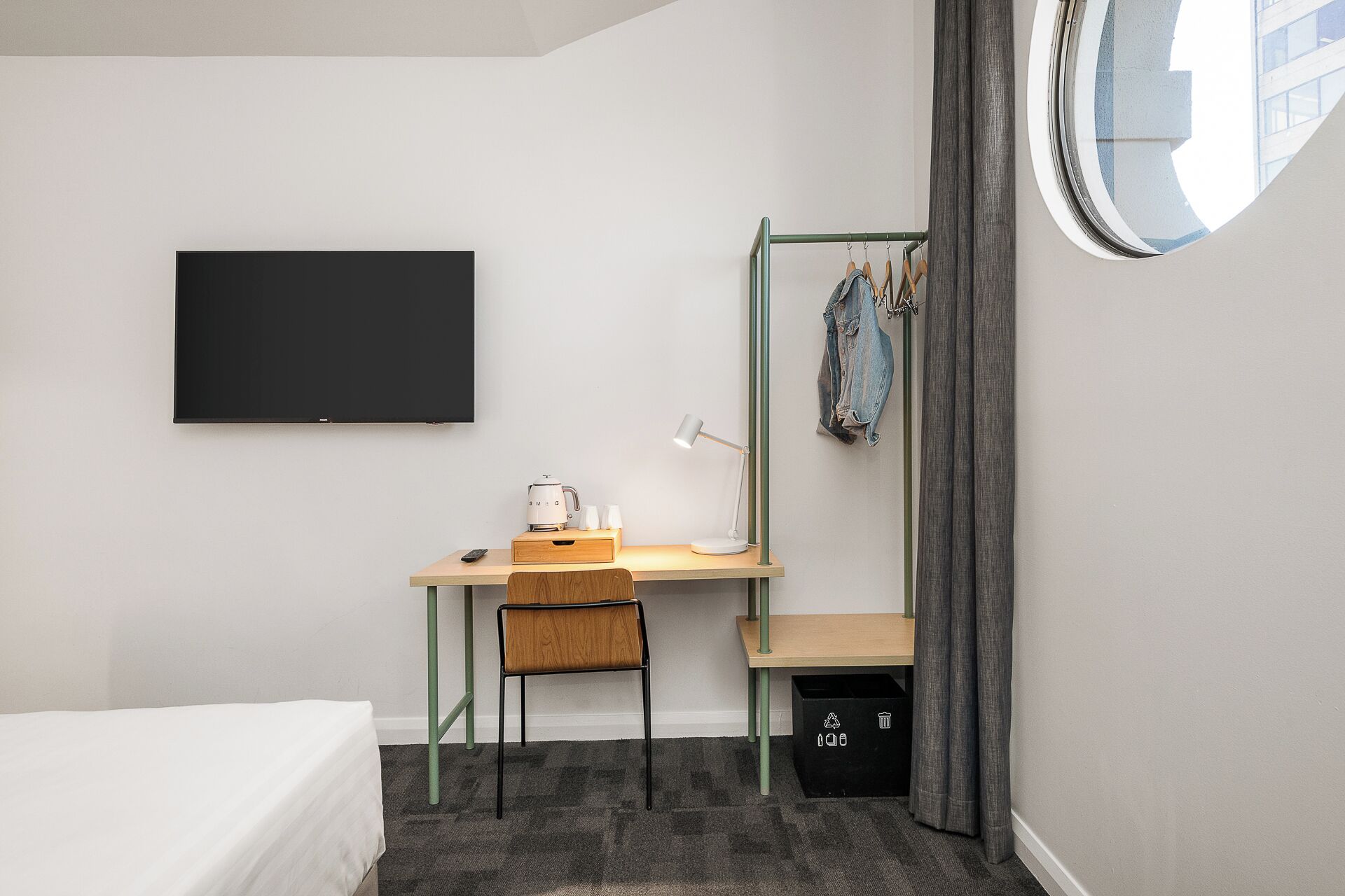 A compact bedroom with a bed, a desk with a chair, a small table with a lamp, a hanging coat rack, and a black trash bin.