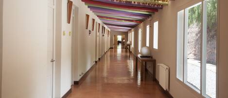 A long, narrow hallway with wooden floors and white walls, featuring a series of doors on one side and large windows on the other.