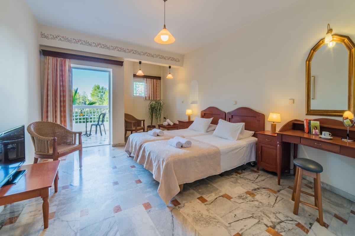 A hotel room with a large bed, two bedside tables, a desk with a mirror, a chair, and a balcony with a view of greenery.