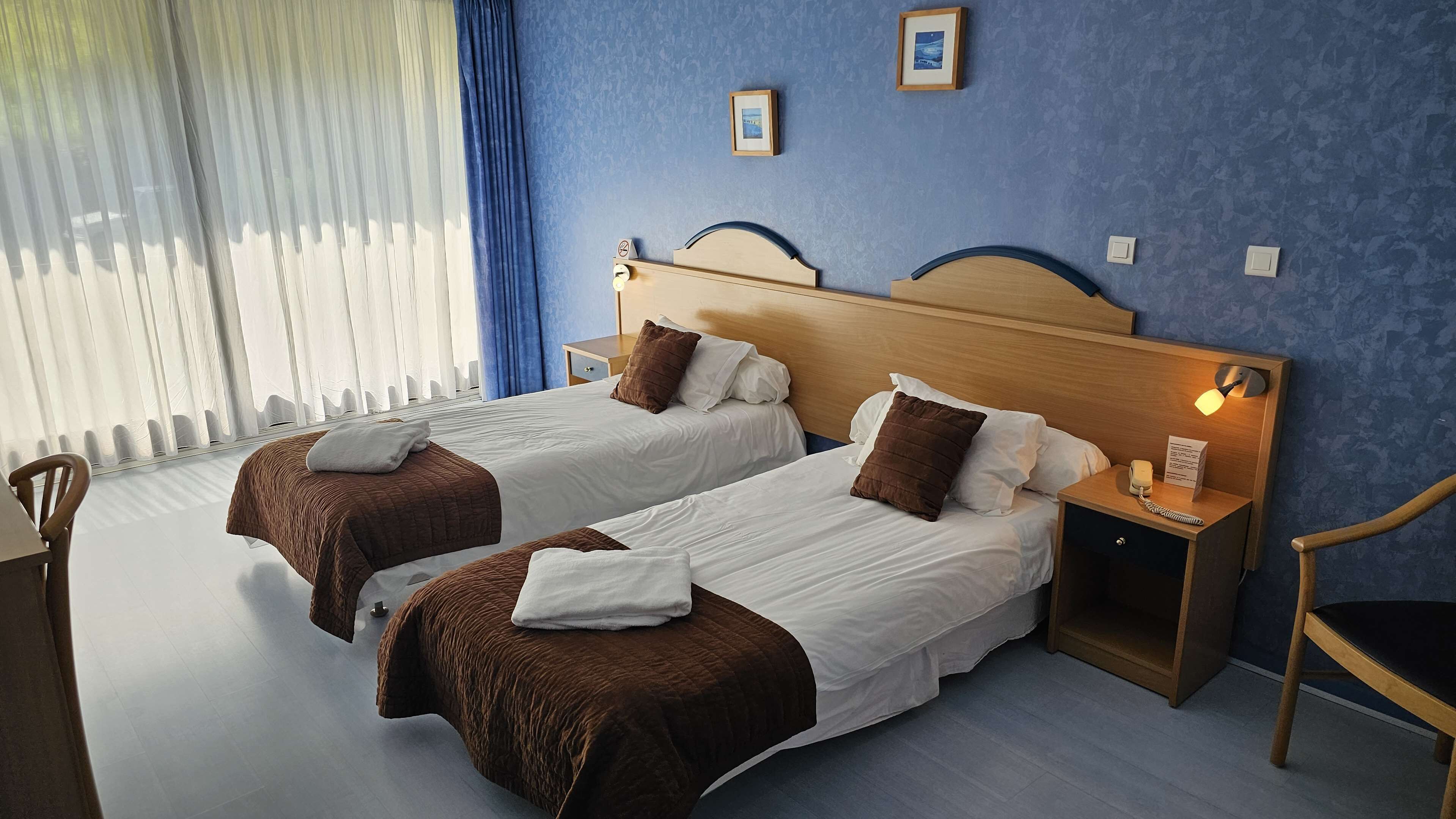 Une chambre d’hôtel avec deux lits, une table de chevet avec une lampe, une chaise et une fenêtre avec des rideaux.