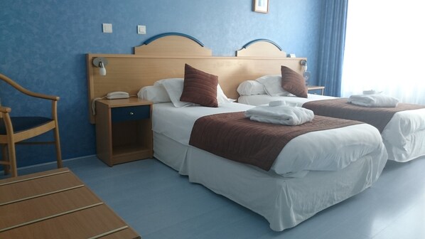Habitación de hotel con dos camas, una mesita de noche de madera con un teléfono, una silla y una pared azul.