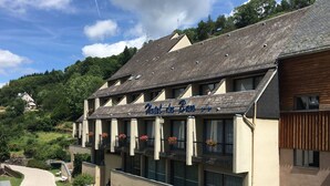 Un hôtel avec une enseigne indiquant « Hôtel du Ban ». Il est doté de nombreux balcons ornés de jardinières fleuries, d’une allée sinueuse et est situé dans un cadre vallonné.