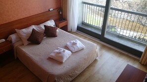 Habitación de hotel con una cama grande, cabecera de madera, dos mesitas de noche y una ventana con vistas a un jardín.