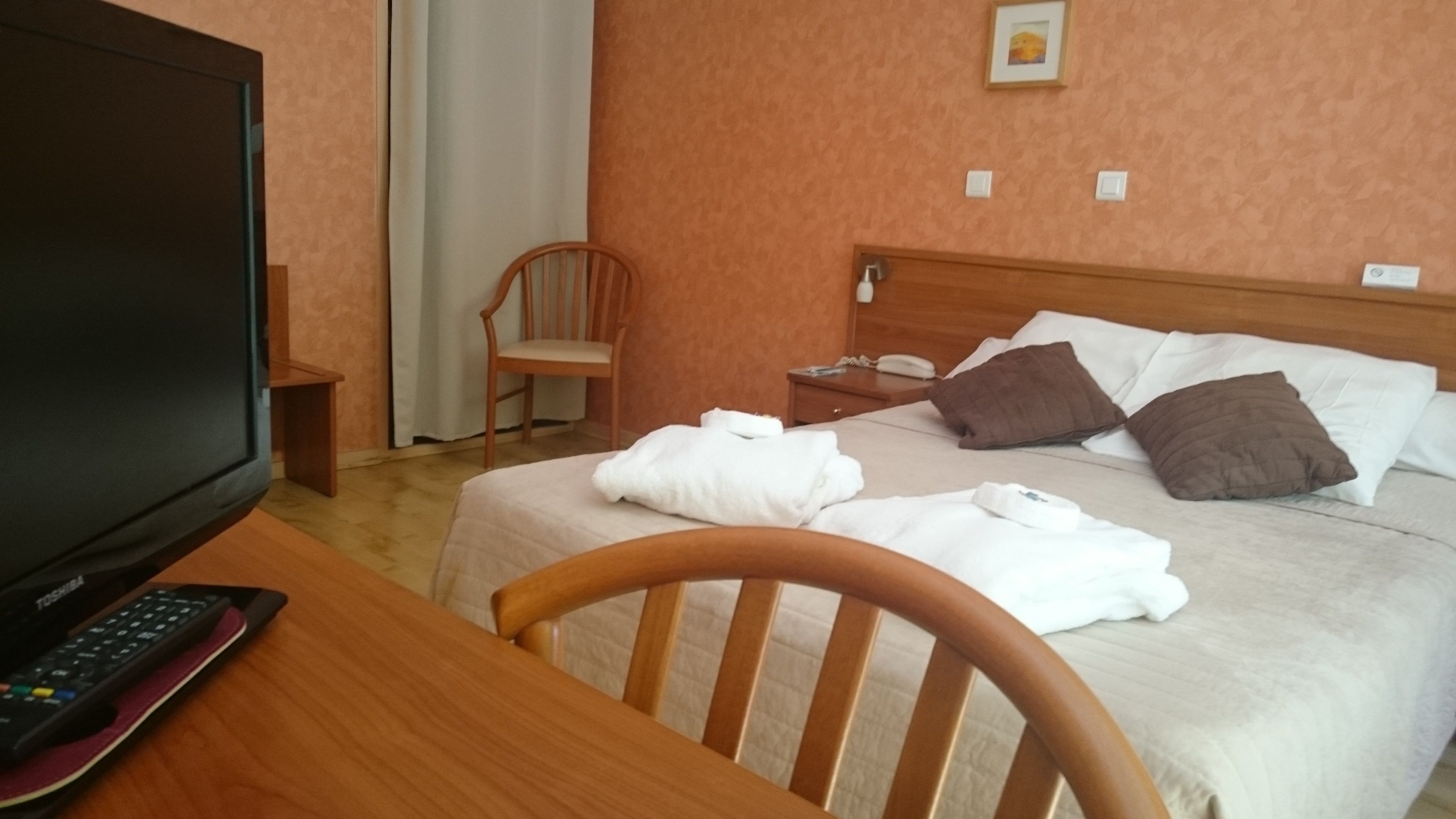 Una habitación de hotel con cama, una silla, un escritorio con televisor y control remoto, una mesita de noche y un cuadro en la pared.
