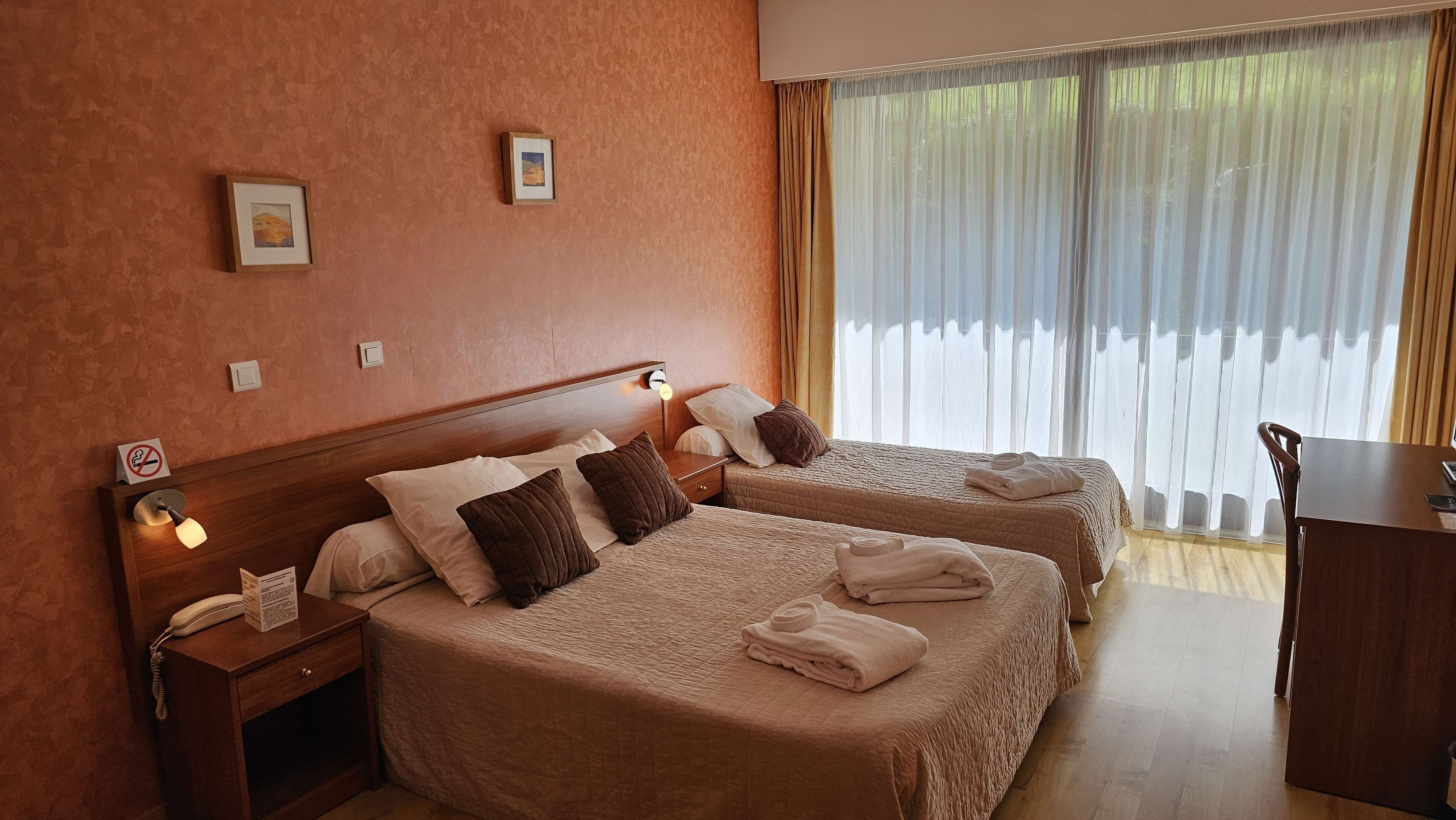A hotel room with two beds, a desk, and a chair. There are two bedside tables with lamps and a phone. The room has a window with curtains.