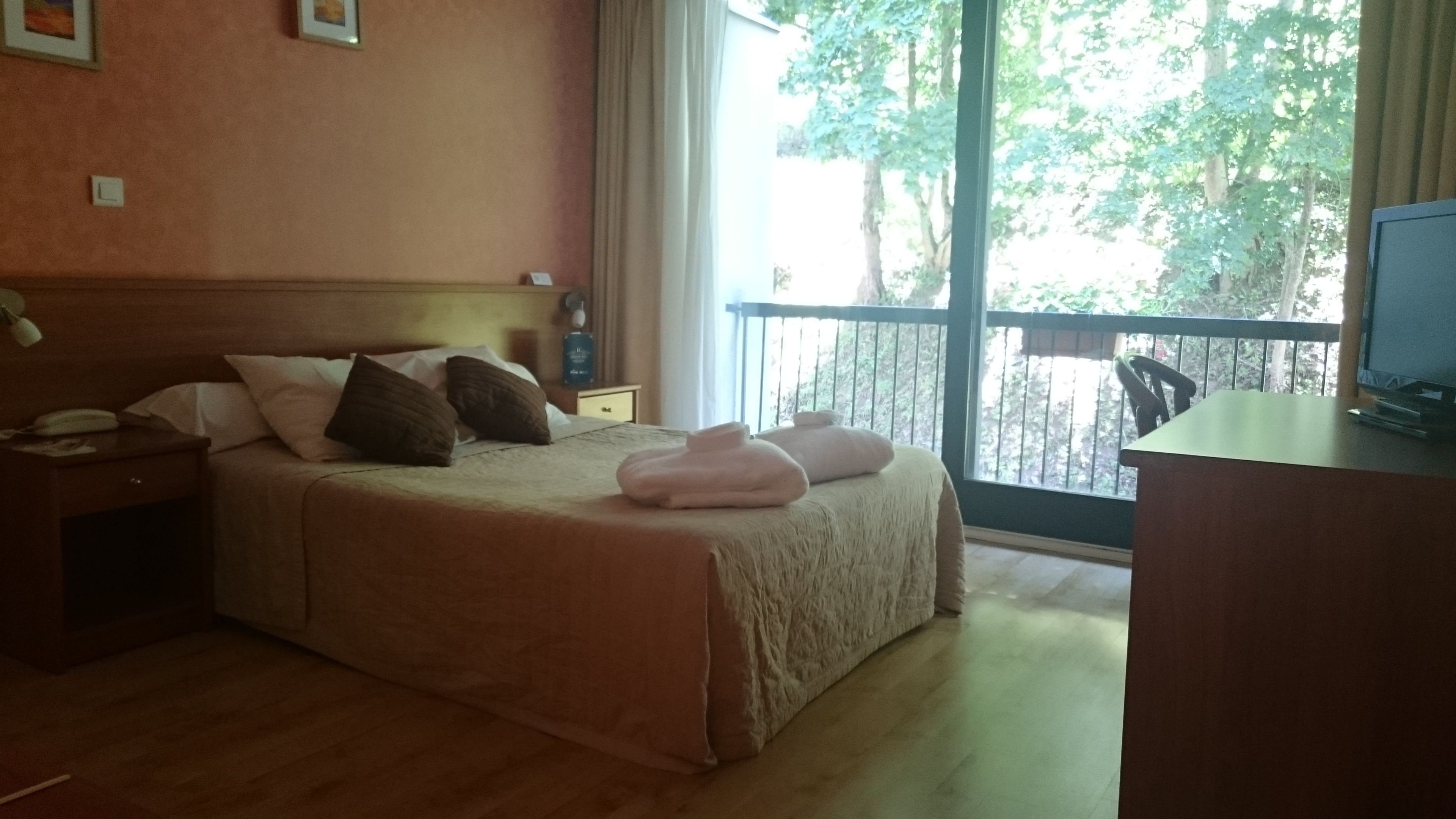 Habitación de hotel con una cama grande, un escritorio con computadora y un balcón con vista a árboles.