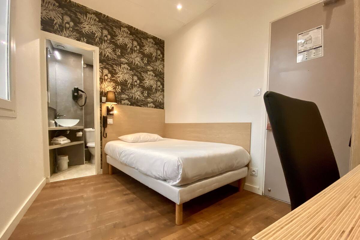 Ein kleines, ordentlich eingerichtetes Schlafzimmer mit einem Einzelbett, einem Nachttisch, einem Stuhl und einem Badezimmer mit Waschbecken und Spiegel.