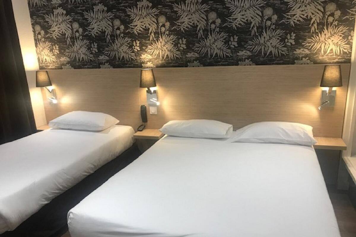 Ein Hotelzimmer mit zwei Einzelbetten, jeweils mit weißer Bettdecke und Kissen, flankiert von Nachttischen mit Lampen und Telefon. An der Wand hinter den Betten ist ein Blumenmuster zu sehen.