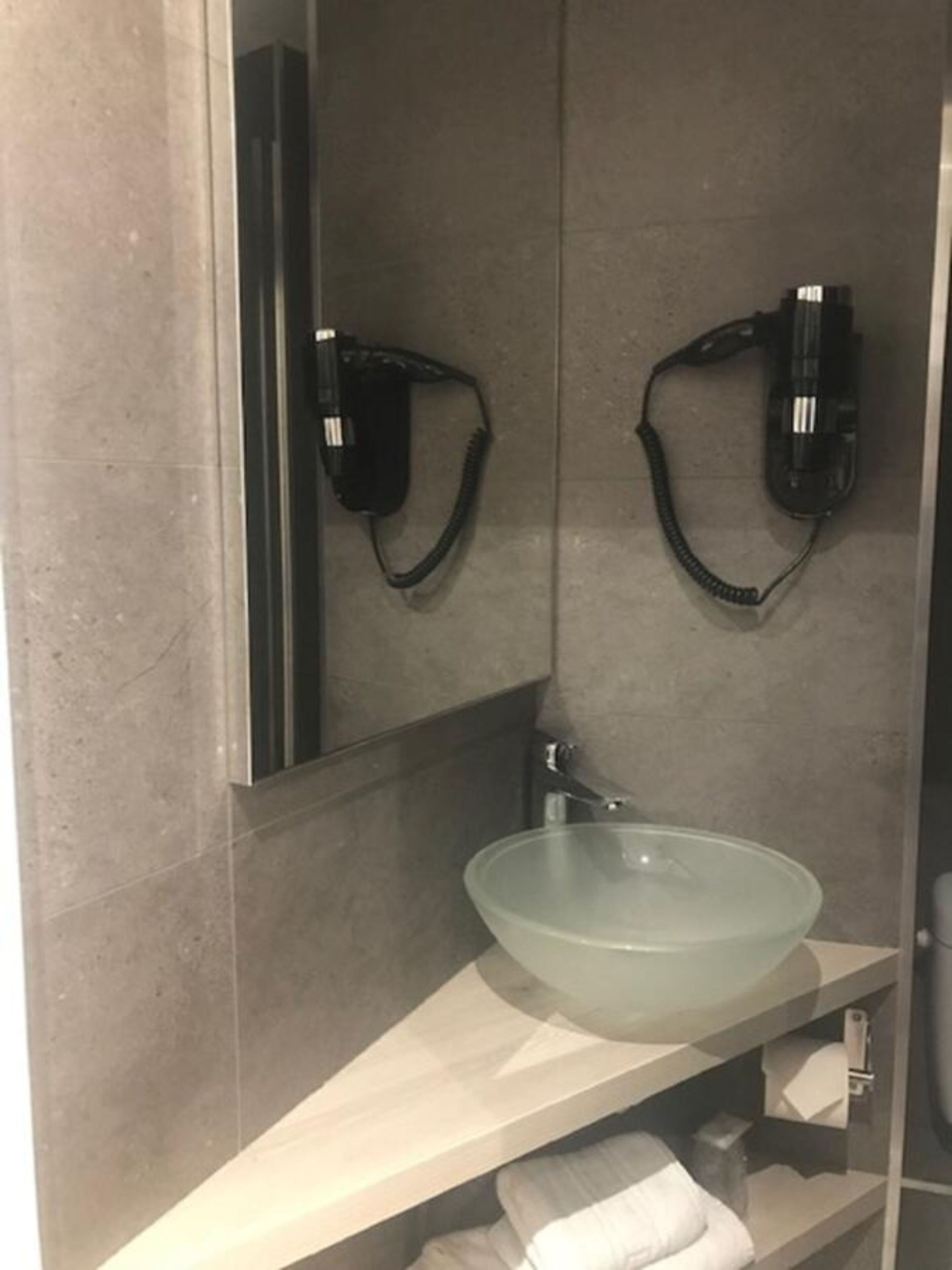 A bathroom with a glass sink, a mirror, and two hair dryers mounted on the wall.
