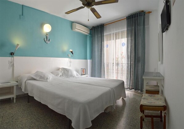 Une chambre d’hôtel avec deux lits, un ventilateur de plafond, un appareil de climatisation, une petite table avec une chaise et un téléviseur fixé au mur.