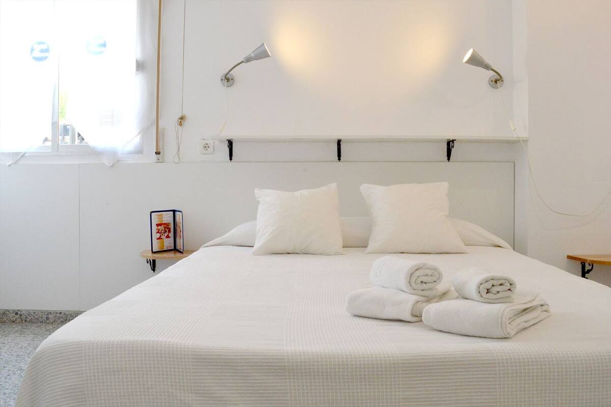 A neatly made bed with white linens and pillows, a bedside table with a framed picture, and two wall-mounted lamps.