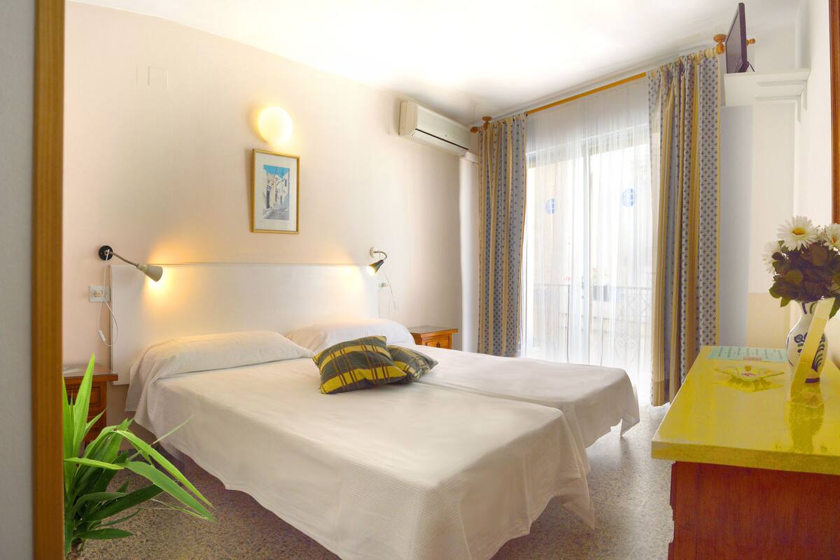 A hotel room with two beds, a bedside table, a desk, and a window with curtains.