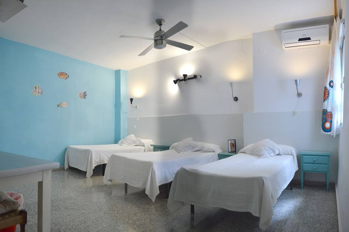 A room with three single beds, a ceiling fan, wall-mounted lights, and a window with a curtain.