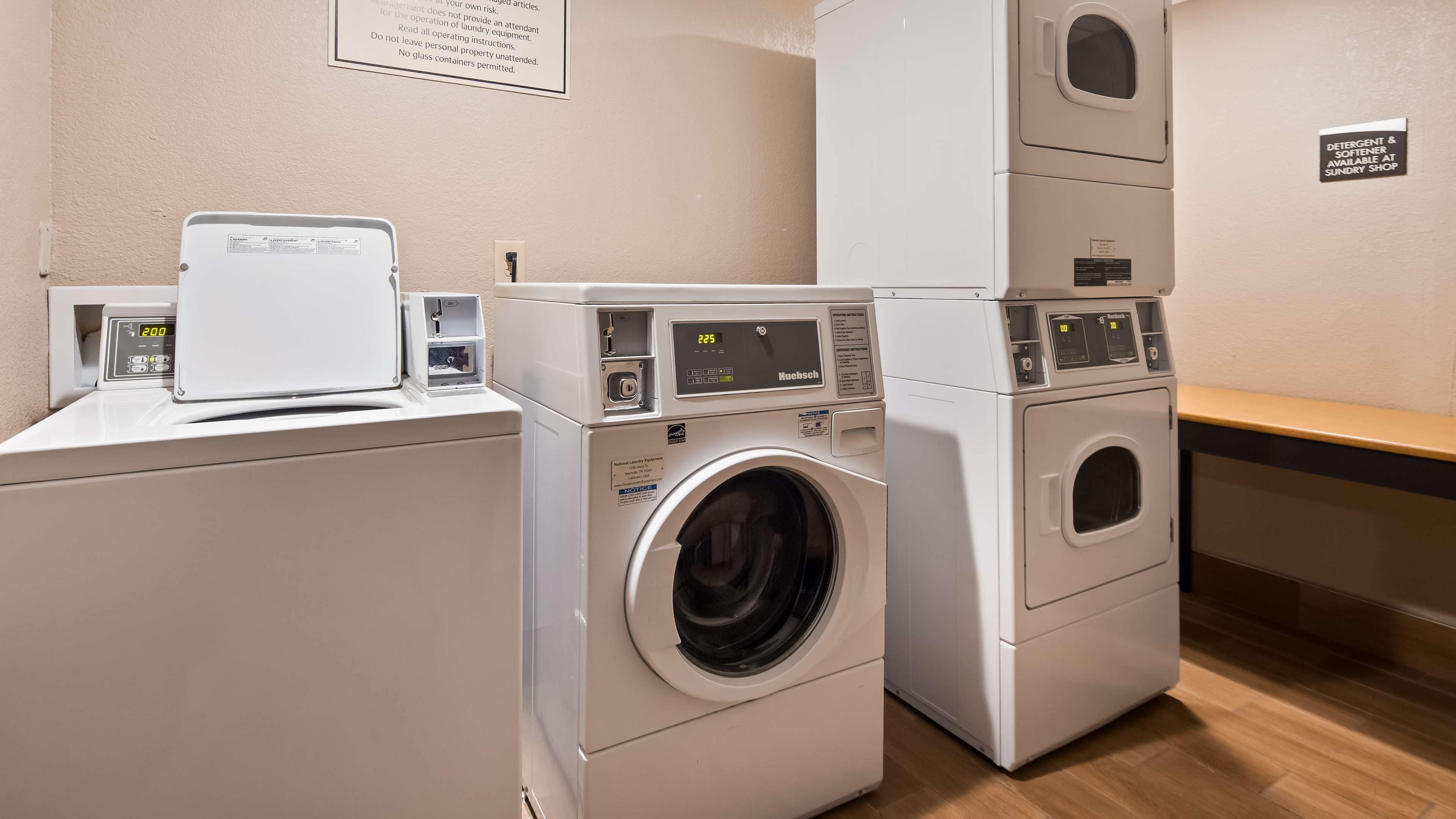 A laundry facility with stacked washers and dryers, a control panel, and a sign indicating detergent and softener availability at the sundry shop.