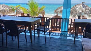 Una terraza de madera con mesas y sillas da a una playa con sombrillas de paja.