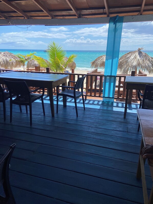 A wooden deck with tables and chairs overlooking a beach with thatched umbrellas.
