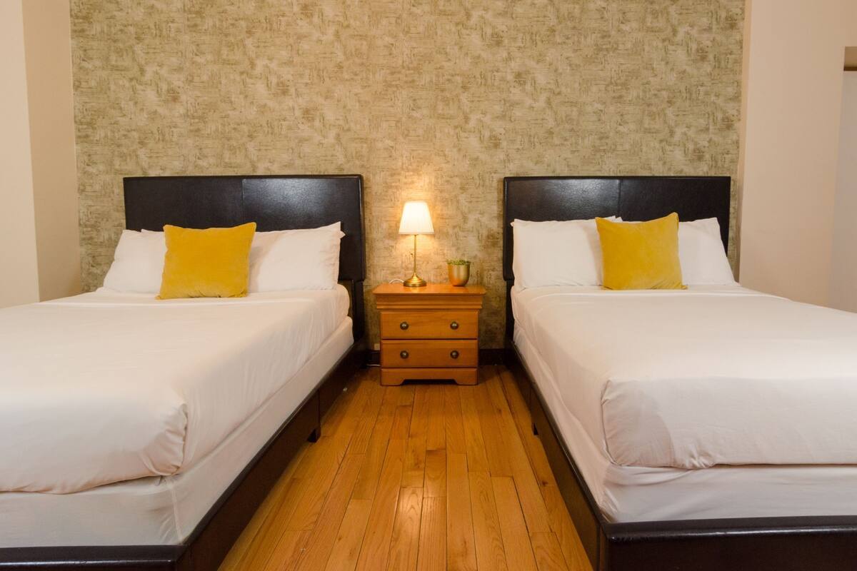 Une chambre d’hôtel avec deux lits, chacun étant équipé de draps blancs et d’un oreiller jaune. Une table de chevet en bois avec une lampe se trouve entre les deux lits.