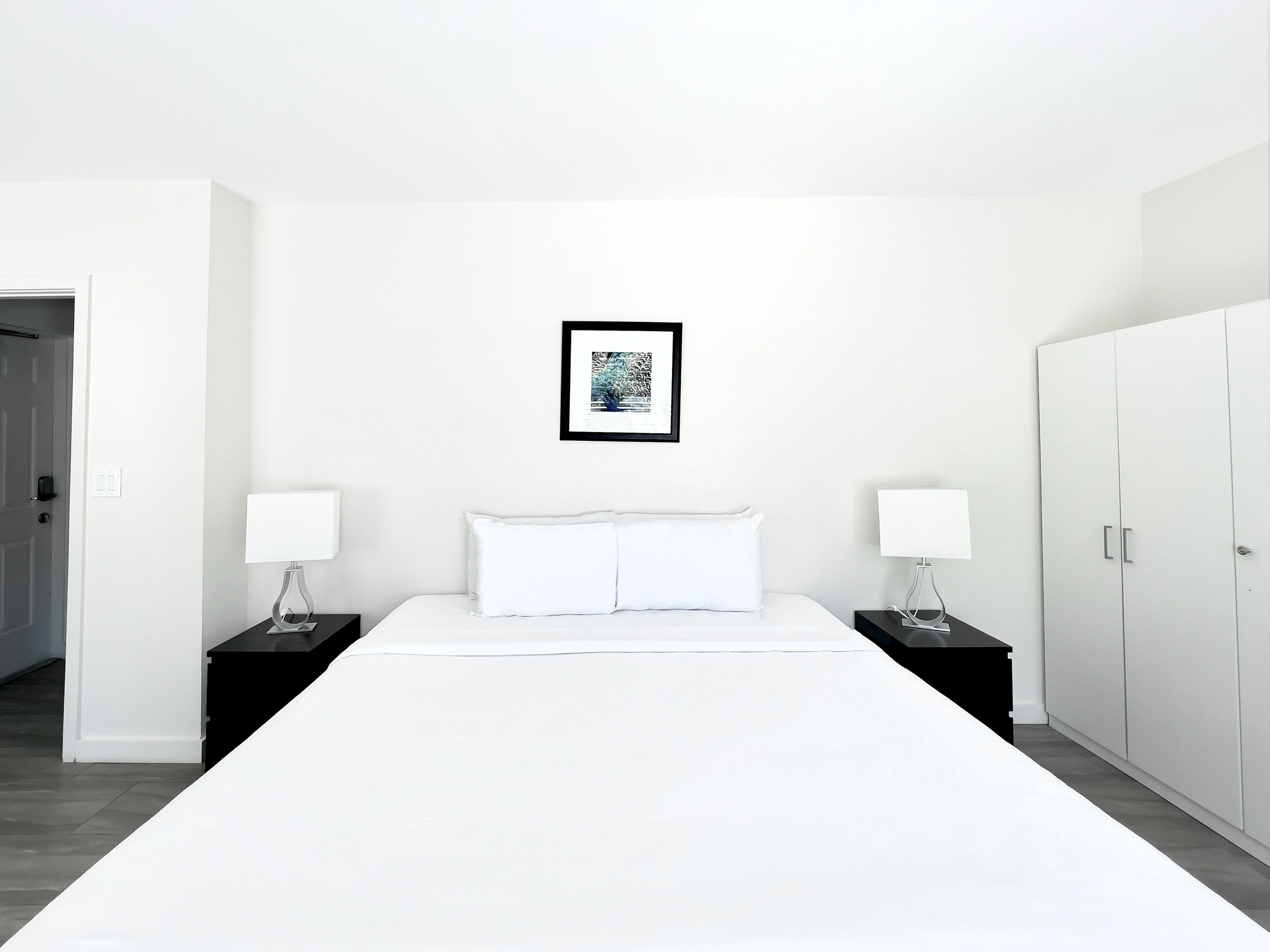 A minimalist hotel room with a large bed, two bedside tables with lamps, a wardrobe, and a framed picture on the wall.